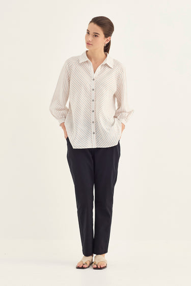A woman modeling a cream-colored long-sleeve button-up shirt with thin vertical gray stripes, paired with dark navy pants by Verge and flat sandals, against a plain white studio background.