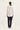 A woman viewed from the back wearing a cream-colored long-sleeve blouse with a fine diagonal striped pattern and dark navy pants by Verge, standing against a plain off-white studio background.