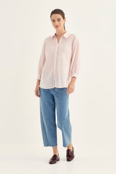 A woman modeling a light pink and white vertically striped button-down shirt with blue cropped jeans by Verge and dark brown loafers, standing against a plain off-white studio background.