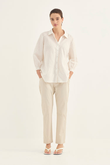 A woman modeling a cream-colored textured button-up shirt and matching beige trousers by Verge against a plain white studio background.
