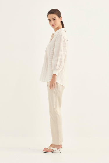 A woman modeling a matching cream-colored two-piece set consisting of a textured long-sleeve button-up shirt and straight-leg trousers by Verge, paired with white heeled sandals, against a plain white studio background.