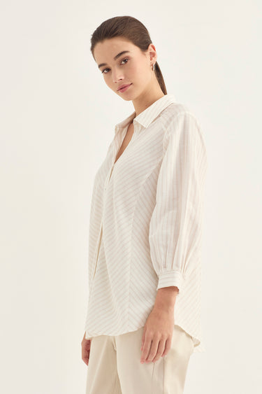 A woman modeling a cream-colored, long-sleeved button-up shirt with thin vertical stripes, paired with matching cream trousers by Verge, against a plain off-white studio background.