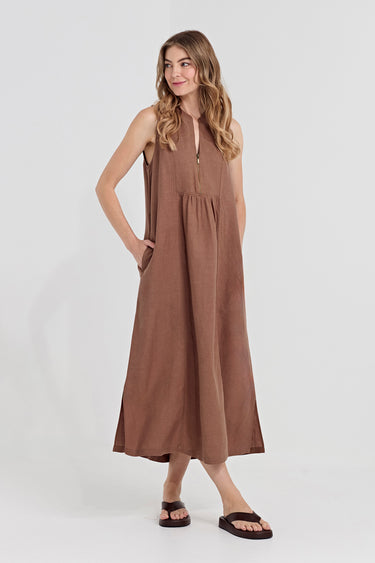 A woman modeling a sleeveless, high-waisted brown maxi dress by Verge with a split neckline and side pockets, paired with dark brown slide sandals, against a light gray studio background.