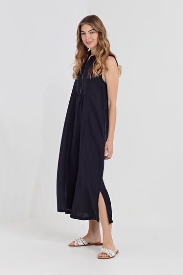 A woman modeling a sleeveless navy blue maxi dress by Verge with a high side slit, paired with white slide sandals, against a plain light gray studio background.