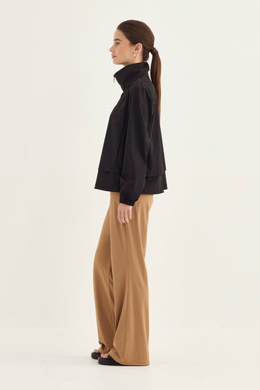 A woman shown in side profile wearing a black long-sleeve high-neck top and wide-leg tan pants by Verge, against a plain light background.