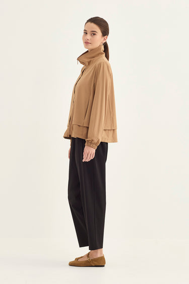 A woman stands in profile wearing a tan cropped jacket with a high collar, black wide-leg cropped pants by Verge, and tan loafers against a plain light background.