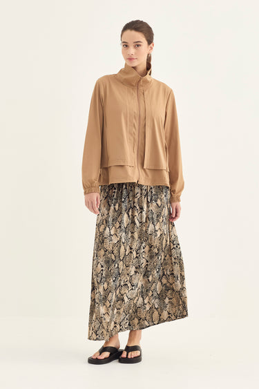 A model stands against a plain light background wearing a tan, cropped zip-front jacket with long sleeves and a high collar, paired with a long skirt by Verge. featuring a beige, brown, and gray snakeskin pattern, and black slide sandals