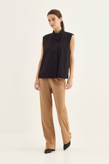 A woman modeling a sleeveless black button-front vest with a high mock neck, paired with tan wide-leg trousers by Verge and black pointed shoes, against a plain off-white studio background.