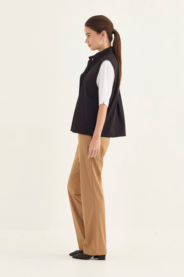 A woman stands in profile wearing a black sleeveless vest over a white short-sleeve shirt, paired with tan wide-leg pants by Verge and black flats, against a plain off-white studio background.