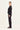 A woman stands in profile wearing a black long-sleeve top with thin vertical striped details on the upper sleeves and black wide-leg pants by Verge, photographed against a plain light background.