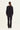 A woman viewed from the back wearing a black long-sleeve top with thin white contrast piping and black trousers by Verge, standing against a plain white studio background.