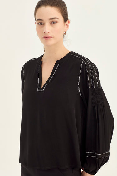 A woman wearing a black long-sleeve blouse by Verge with decorative white embroidered details along the split neckline, shoulders, and cuffs, photographed against a plain light background.