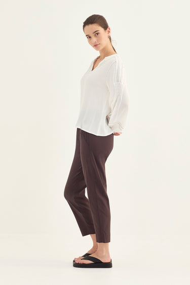 A woman modeling a white long-sleeve blouse and dark brown cropped pants by Verge with black sandals, standing against a plain white studio background.