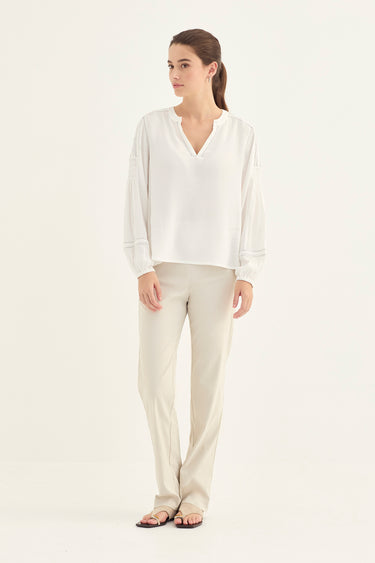 A woman modeling a white long-sleeve V-neck blouse and light beige straight-leg pants by Verge against a plain white studio background.