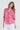 A woman modeling a vibrant pink and white patterned blouse with three-quarter sleeves, paired with white pants by Verge, against a plain light gray background.