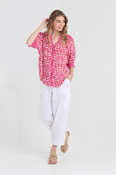 A woman modeling a short-sleeve button-up blouse with a vibrant pink and white leaf pattern, paired with white cropped trousers by Verge and beige sandals, set against a plain white studio background.