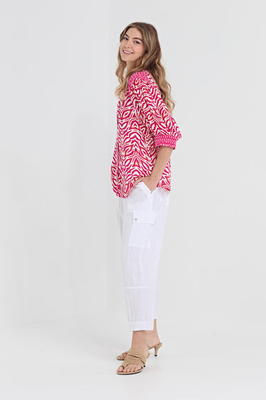 A woman modeling a pink and white patterned short-sleeve top paired with white pants by Verge and beige heeled sandals, standing against a plain light gray studio background.