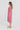 A woman stands in profile wearing a loose, short-sleeve midi dress by Verge with an allover pink and white abstract wavy pattern, paired with white slide sandals, against a plain light gray studio background.