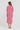 A woman viewed from the back wearing a pink and white patterned midi dress by Verge with short puff sleeves and a gathered waist, paired with flat sandals, against a plain white studio background.