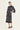 A woman modeling a black midi shirt dress by Verge with white botanical and tribal patterns, paired with black slide sandals, against a plain off-white studio background.