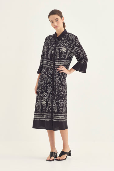 A woman modeling a black midi shirt dress by Verge with white botanical and tribal patterns, paired with black slide sandals, against a plain off-white studio background.