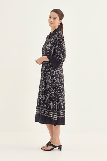 A woman modeling a black dress by Verge with a white floral print, shown in profile against a plain light background.
