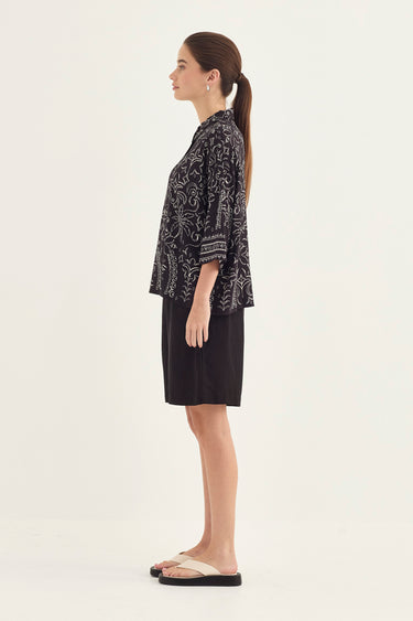 A woman shown in side profile wearing a black and white patterned short-sleeve top and a black skirt by Verge, standing against a plain light background.