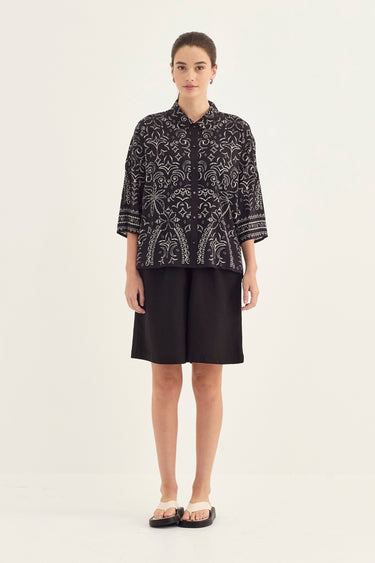 A woman stands against a plain light background wearing a black short-sleeve button-front blouse with an intricate white botanical pattern by Verge, paired with black shorts and black slide sandals.