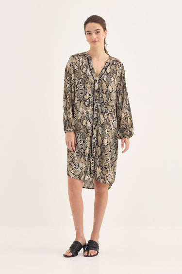 A woman modeling a long-sleeve button-front shirt dress by Verge with a beige, brown, and black snakeskin pattern, paired with black slide sandals, against a plain off-white studio background.
