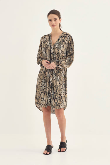 A woman modeling a long-sleeve, knee-length button-front dress by Verge with an allover snakeskin print in beige, brown, and black, paired with black slide sandals, against a plain white studio background.