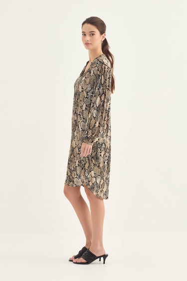 A woman modeling a long-sleeve, knee-length dress by Verge with a beige, brown, and black snakeskin pattern, paired with black mule sandals, set against a plain off-white studio background.