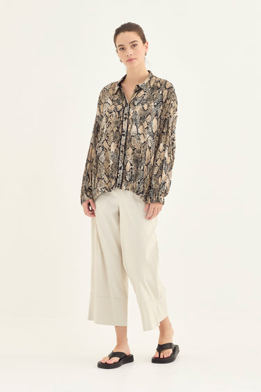 A woman modeling a long-sleeve button-up shirt with a beige, brown, and black snakeskin pattern, paired with cream cropped trousers by Verge and black slide sandals, against a plain off-white studio background.