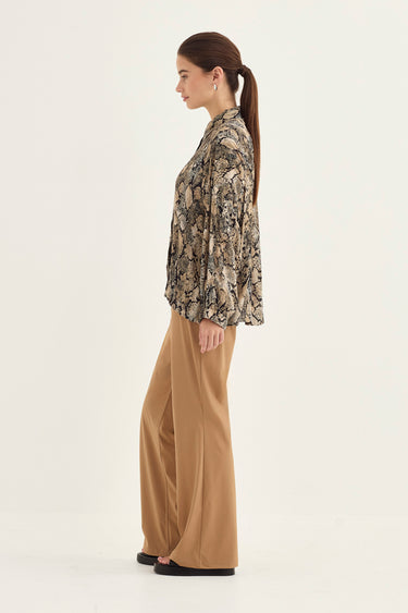 A woman shown in profile wearing a long-sleeve button-up blouse with a beige, brown, and black abstract pattern, paired with wide-leg tan pants by Verge and black flat shoes, against a plain off-white studio background.