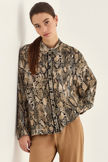A woman wearing a long-sleeve button-up blouse with a beige and black snakeskin pattern, paired with tan trousers by Verge, leaning against a plain off-white wall.