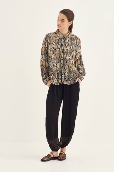 A woman modeling a long-sleeve button-up shirt with a beige and brown snakeskin pattern, paired with black pants by Verge and flat sandals, against a plain white studio background.