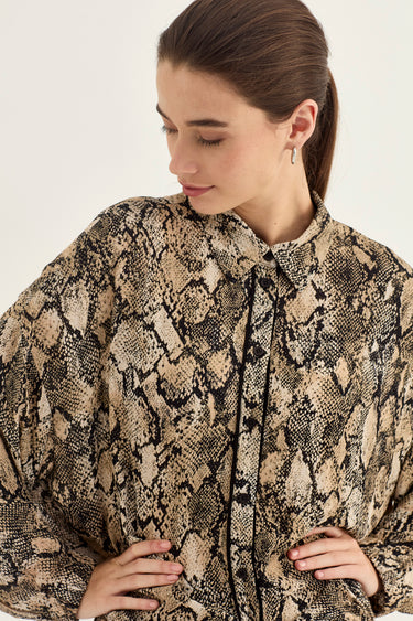 A woman wearing a long-sleeve collared blouse by Verge with a beige, brown, and black snakeskin pattern, photographed against a plain light background.