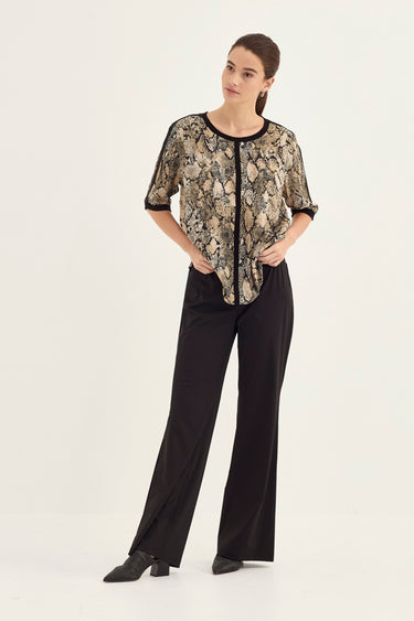 A woman modeling a short-sleeve blouse with a beige and black snakeskin pattern, paired with black wide-leg trousers by Verge, against a plain white studio background.