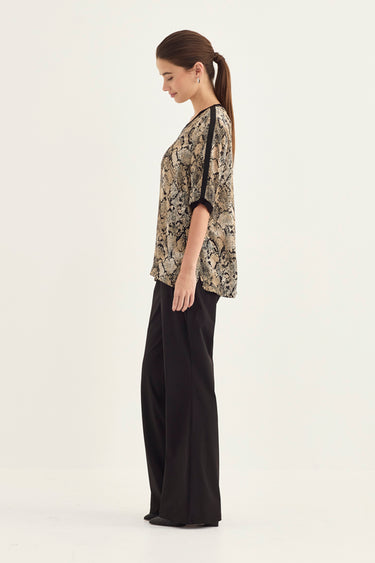 A woman stands in profile wearing a short-sleeve top with a beige, black, and brown abstract pattern and black wide-leg pants by Verge against a plain light background.