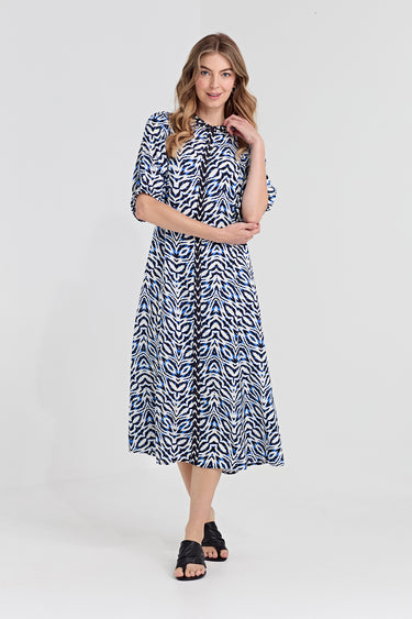 A woman modeling a midi dress by Verge with a blue and white abstract wavy pattern, featuring elbow-length puff sleeves and a gathered waist, paired with black slide sandals against a light gray studio background.
