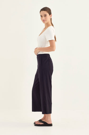 A woman modeling a pair of dark navy wide-leg cropped pants by Verge with a white short-sleeve top and black sandals, shown in profile against a plain white studio background.