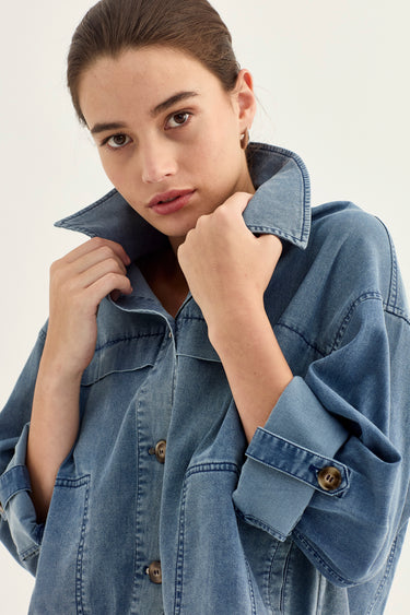 A close-up of a woman wearing an oversized blue denim shirt jacket by Verge with rolled-up sleeves and a collar, posed against a plain light background.