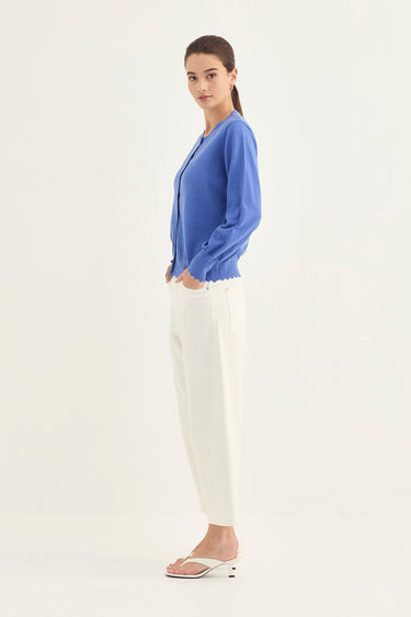 A woman stands in profile wearing a blue long-sleeve cardigan, white pants by Verge, and white heeled sandals against a plain light background.