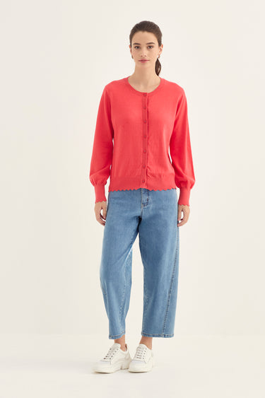 A woman modeling a coral red long-sleeve button-up cardigan with voluminous sleeves, paired with light blue cropped jeans by Verge and white sneakers, against a plain off-white studio background.