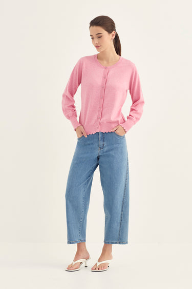 A woman modeling a light pink long-sleeve knit cardigan with a scalloped hem, paired with light blue cropped jeans by Verge and white slide sandals, against a plain off-white studio background.
