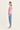 A woman stands in profile wearing a light pink long-sleeve knit top, light blue jeans by Verge, and white heeled sandals against a plain off-white studio background.