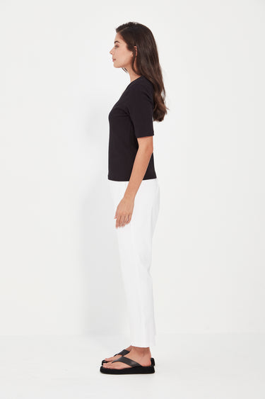 A woman shown in profile wearing a fitted white short-sleeve top, dark navy cropped pants by Verge, and black sandals against a plain light gray studio background.