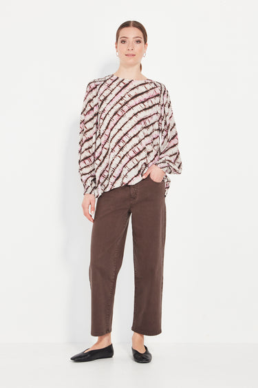 Woman wearing a patterned blouse and brown pants by Verge on a white background