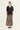 A woman stands against a plain light background wearing a black short-sleeve V-neck top and a long snakeskin print maxi skirt by Verge in beige, brown, and black tones, with black sandals.