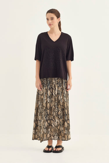 A woman stands against a plain light background wearing a black short-sleeve V-neck top and a long snakeskin print maxi skirt by Verge in beige, brown, and black tones, with black sandals.
