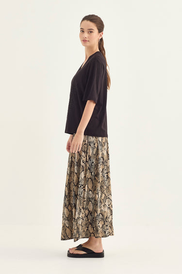 A woman stands in profile wearing a loose black short-sleeve top and a long pleated skirt with a beige, brown, and black snakeskin pattern by Verge, paired with black flip-flops, against a plain off-white studio background.